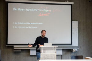 Lukas Kohmann is standing in the middle of the image behind a lectern. He is speaking. In the backround is a powerpoint presentation with the words: The space of Artificial Intelligence