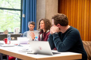 Two men and a woman sitting at a table, with laptops in front of them. The woman is speaking while the men are listening and looking at her.