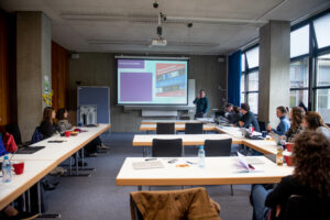 Michael Pelzer ist standing in the centre of the image. He is holding a presentation, while people on the right an left side of the image are sitting at tables and listening.