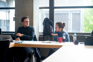 A man on the left and a woman on the right are sitting at a table. They look at each other and discuss something. There is a window in the background.