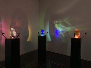 the image shows three steles which each display a oval shaped glass object. Inside of each object there is a colourful glowing material. The one on the left is plum-coloured, the one in the middle indigo and the one on the right orange.