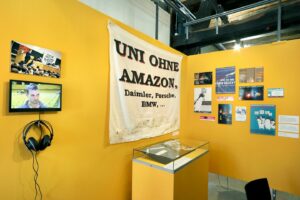 The Image shows a corner of the exhibition with yellow walls. There are various posters and transparents on the wall. On the left there is a big poster which says: "Uni ohne Amazon" in capital letters.

Image: Anne Faden