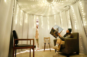 The image shows the inside of a white pavillon. There are fairy lights hanging in a star-shaped construction from the ceiling of the pavillon. There are three seats on the bottom of the image. One on the left, one in the middle and one on the right. On the right seat, there is a young woman sitting on it and holding a magazine. On the middle.left side of the image there is another person visible. The person is peaking throug a little opening inside the Pavillon.

Image: Anne Faden