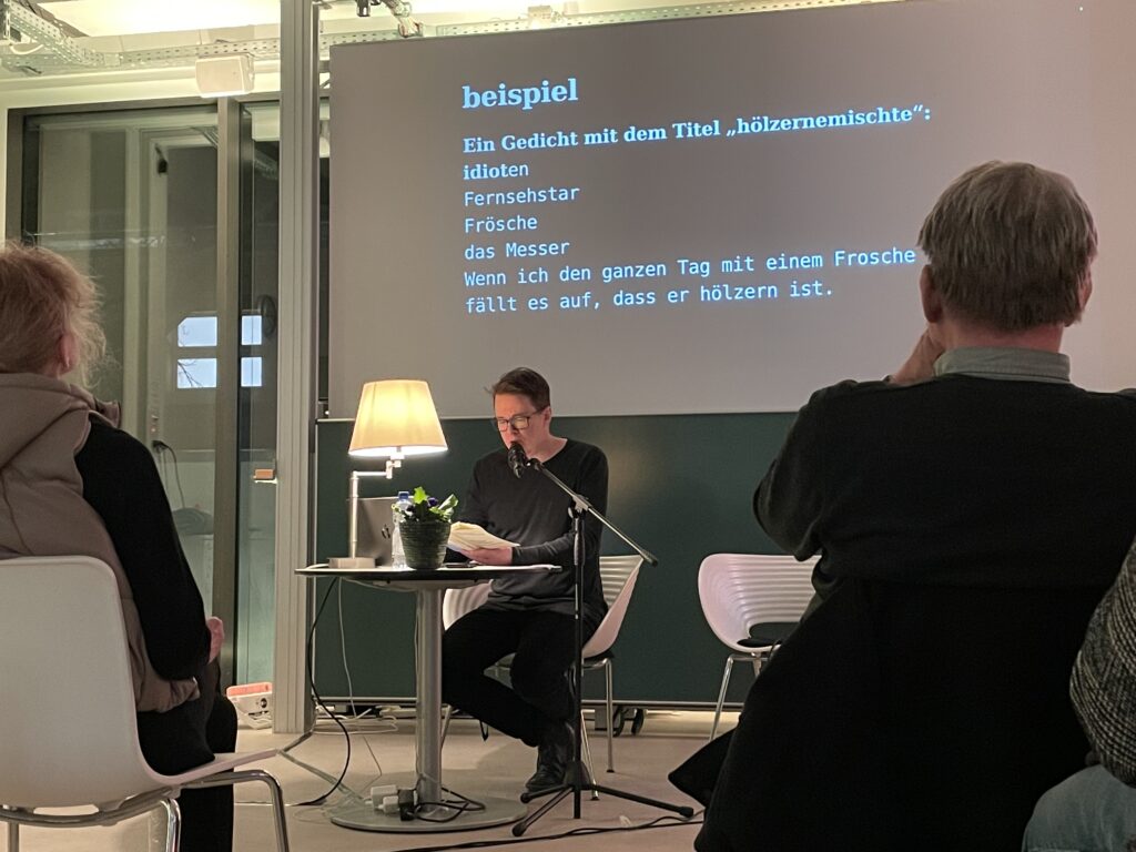 Jörg Piringer is sitting in the centre of the image. He is reading from his book. In the background you can see a projection of the poem he is reading at the moment.