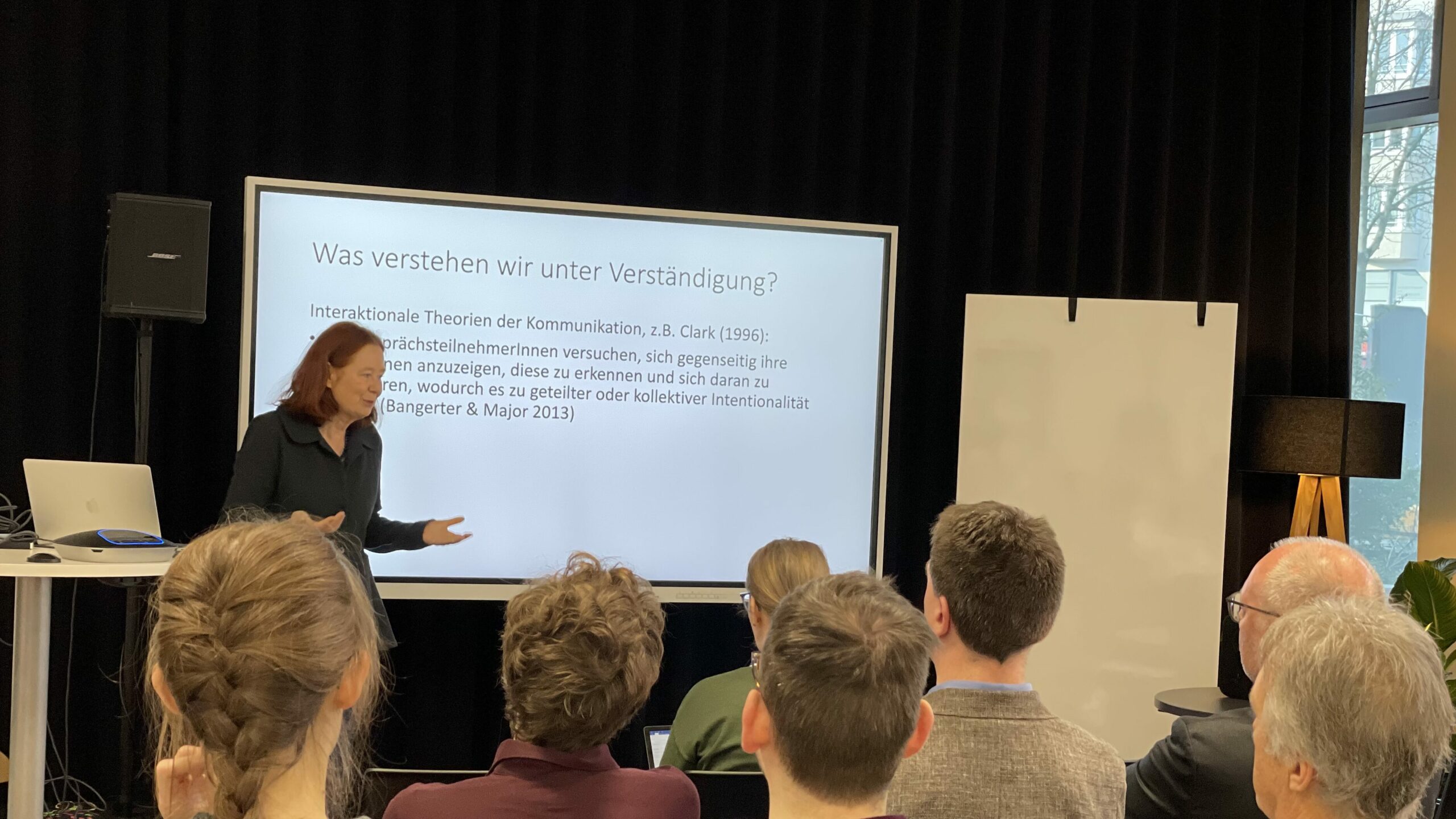 Professor Kerstin Fischer steht vor einer Präsentationsfolie auf der steht "Was verstehen wir unter Verständigung? Interaktionale Theorien der Kommunikation, z.B. Clark (1996)".