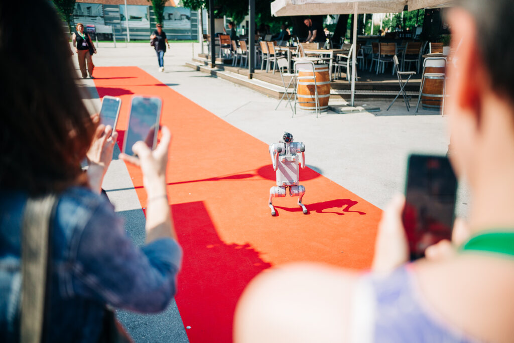 Ein langer roter Teppich, im Hintergrund Menschen und der Außenbereich eines Restaurants; auf dem Teppich ein weißer Roboter, im Vordergrund unscharf mehrere Menschen, die mit Smartphones Aufnahmen von dem Roboter machen. 