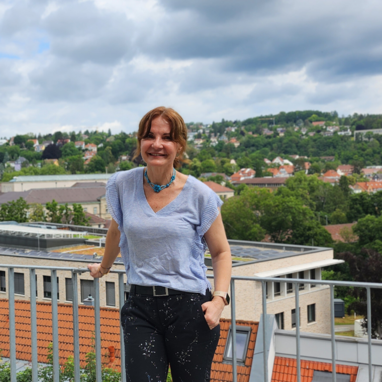 Maria lächelt in die Kamera. Sie steht draußen und lehnt sich an ein Geländer. Im Hintergrund ist ein Ausblick über Tübingen mit Universitätsgebäuden und grünen Bäumen zu sehen.