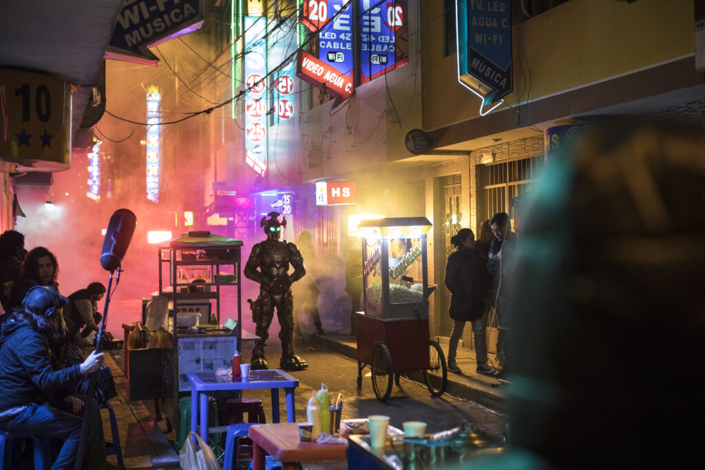 The image shows the scenery of a city by night. In the middle of well-lit shops and street vendors there is a humanoid lokking poboter with bright green eyes.