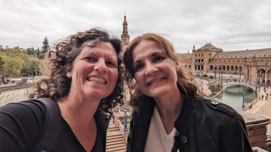 Das Bild ist ein Selfie von Maria del Mar und Anne Burkhardt. Sie lächeln in die Kamera. Im Hintergrund ist die Architektur Sevillas zu sehen.