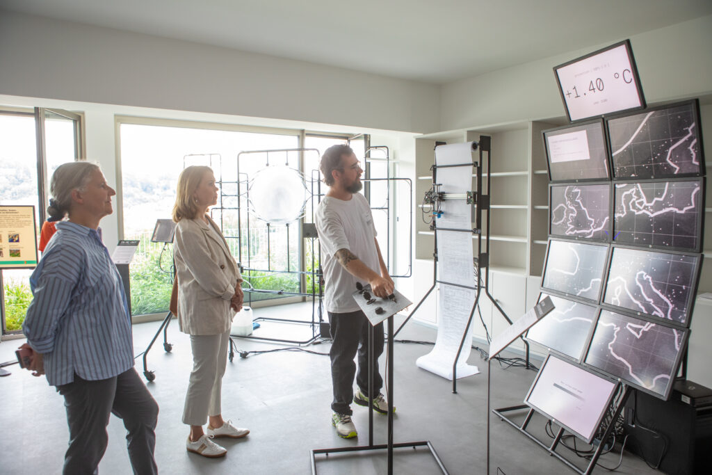 The image shows Karla Pollmann and Susanne Olschowski in the Showroom looking at the IN-ML-Out- artwork. A man in a white shirt explains the artwork to them.