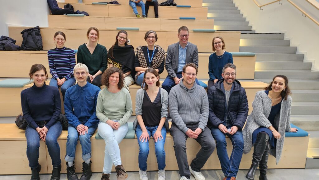 Gruppenfoto von 13 Menschen, die auf Stufen wie in einem Atrium sitzen.