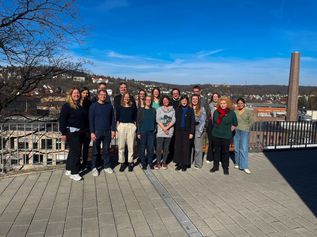 Gruppenfoto mit 17 Menschen, die draußen auf einem gepflasterten Hof stehen: Im Hintergrund blauer Himmel und der Campus der Stadt Tübingen.
