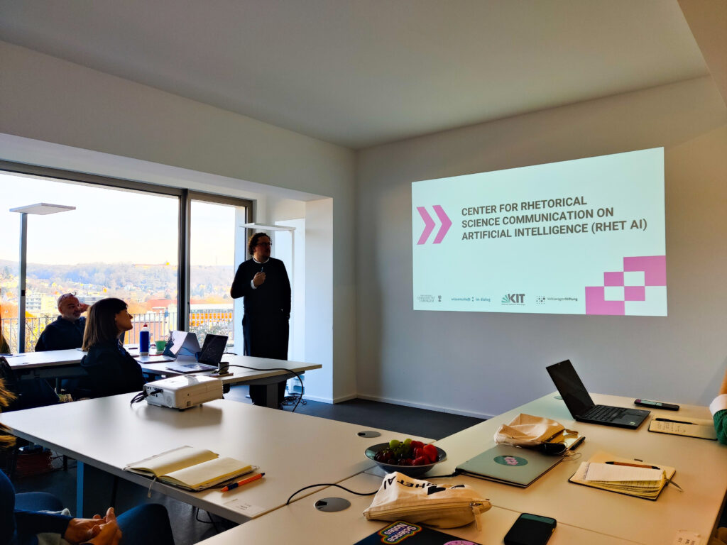 Seminarraum mit mehreren Tischen, Laptops, Schreibutensilien und Beamer und zwei zuhörende Personen im Vordergrund. Im Hintergrund eine Person, die eine Präsentation hält, an die Wand projiziert ein Slide einer Präsentation.