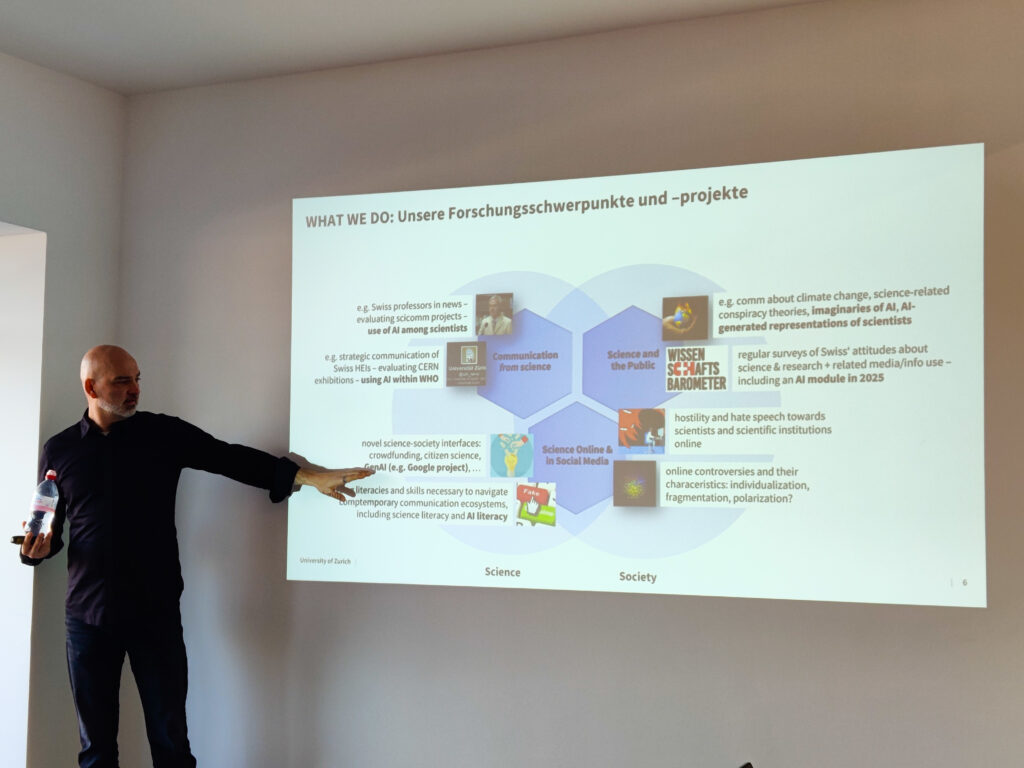 Im Vordergrund ein Mann in dunkler Kleidung mit einer Wasserflasche in der Hand. Er redet und deutet gleichzeitig auf das Slide einer Präsentation im Hintergrund. Dort sind mehrere Textblöcke und Grafiken zu sehen.