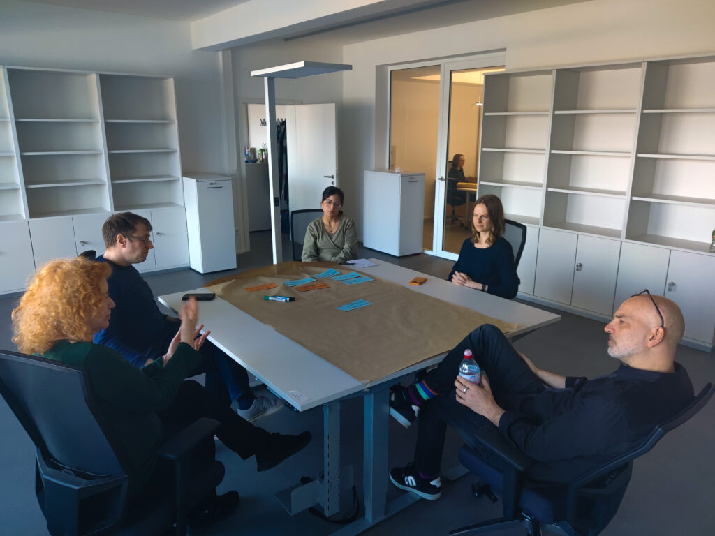 5 Menschen sitzen in einem Büroraum um einen Tisch und unterhalten sich. In der Mitte liegen ein großes Papierplakat und Schreibtuensilien.