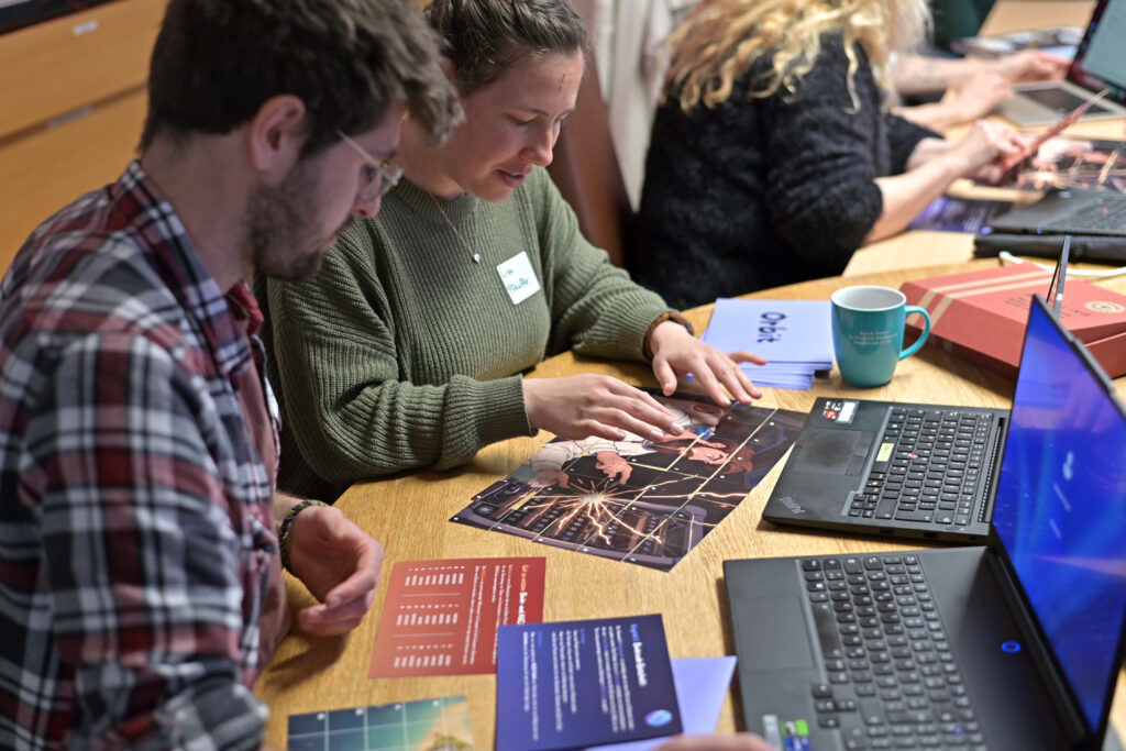 Zwei Personen haben Laptops vor sich und fügen auf einem Tisch bunte Puzzleteile zusammen. Im Hintergrund sitzt eine weitere Person am Tisch und arbeitet.