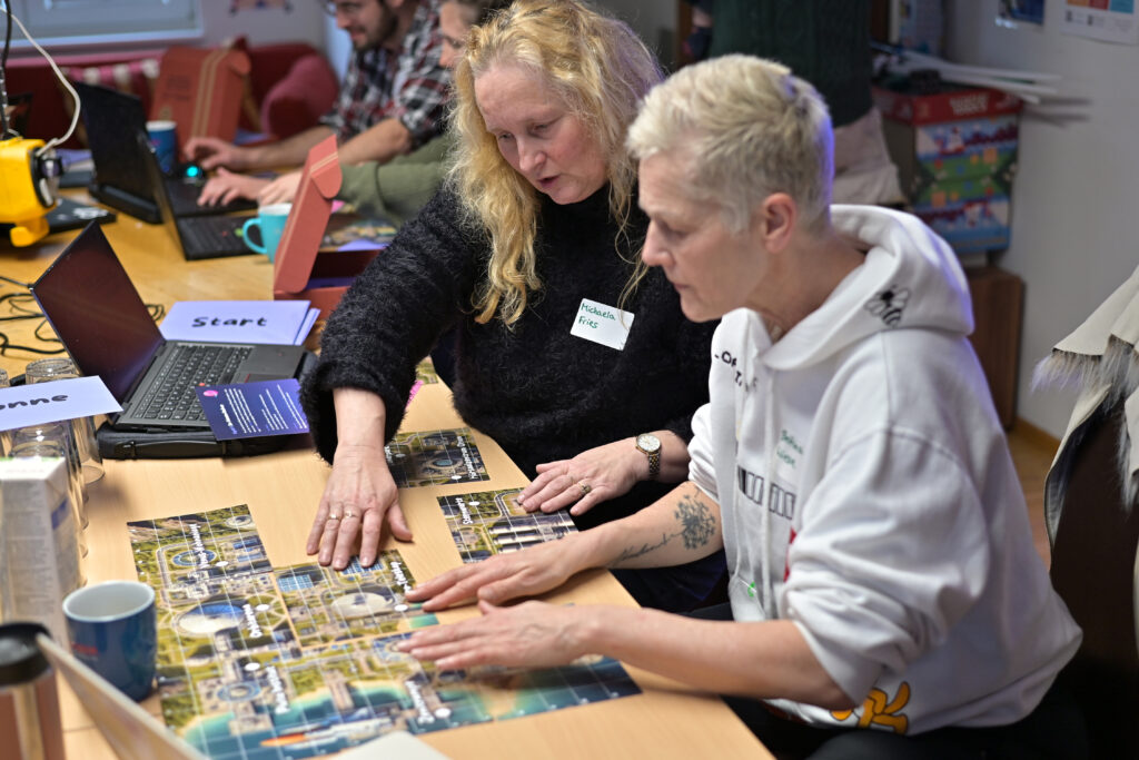 Zwei Personen fügen bunte Karten zu einem Puzzle zusammen und schauen konzentriert. Auf dem Tisch stehen Laptops und Tassen, im Hintergrund arbeiten weitere Menschen am Laptop.