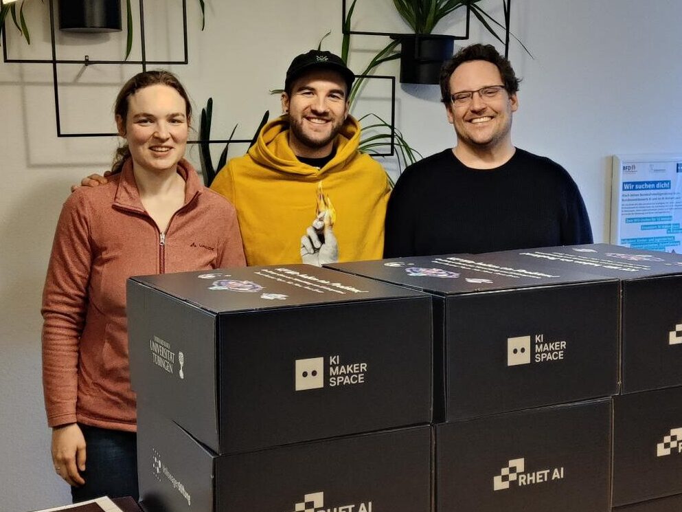 Drei Personen schauen fröhlich in die Kamera, vor ihnen ein großer Stapel von Boxen mit dem RHET AI Logo und dem Logo des KI-Makerspace Tübingen.