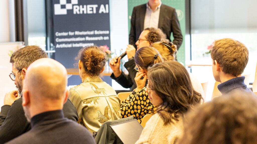 mehrere Menschen sitzen in einem Tagungsraum und hören einem Vortrag zu, einer hält ein Mikrofon und spricht. Im Hintergrund steht eine weitere Person. Links im Hintergrund ist ein Banner mit weißer Schrift auf dunklem Grund mit dem RHET AI Logo.