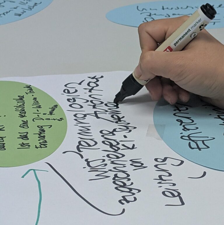 Eine Hand schreibt mit einem dicken schwarzen Filzstift in großen Buchstaben auf ein Blatt Papier. Daneben liegen mehrere kleinere beschriebene Paperikarten in blau und grün.