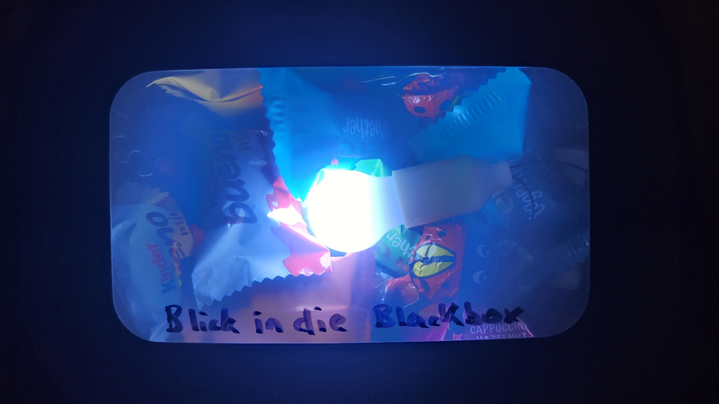 Das Bild zeigt eine blau-lila leuchtende, metallene Brotdose vor schwarzem Hintergrund. Auf der Brotdose ist "Für die BlackBox" geschrieben.