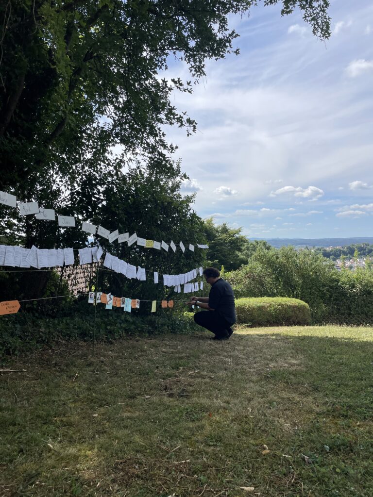 Das Bild zeigt eine Wäscheleine voller Zettel, die in einem Garten gespannt ist. Vor der Wäscheleine kniet ein Mann.