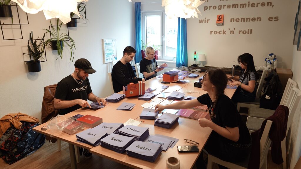 Das Bild zeigt fünf Menschen, die an einem großen Tisch sitzen und Materialien für das KI- Escape Game einpacken.