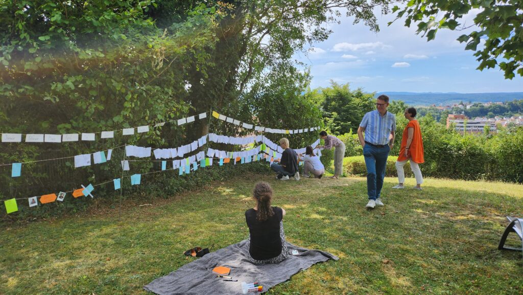 Das Bild zeigt eine Wäscheleine voller Zettel, die in einem Garten gespannt ist. Im Vordergrund sind Menschen zu sehen, die die Wäscheleine betrachten.
