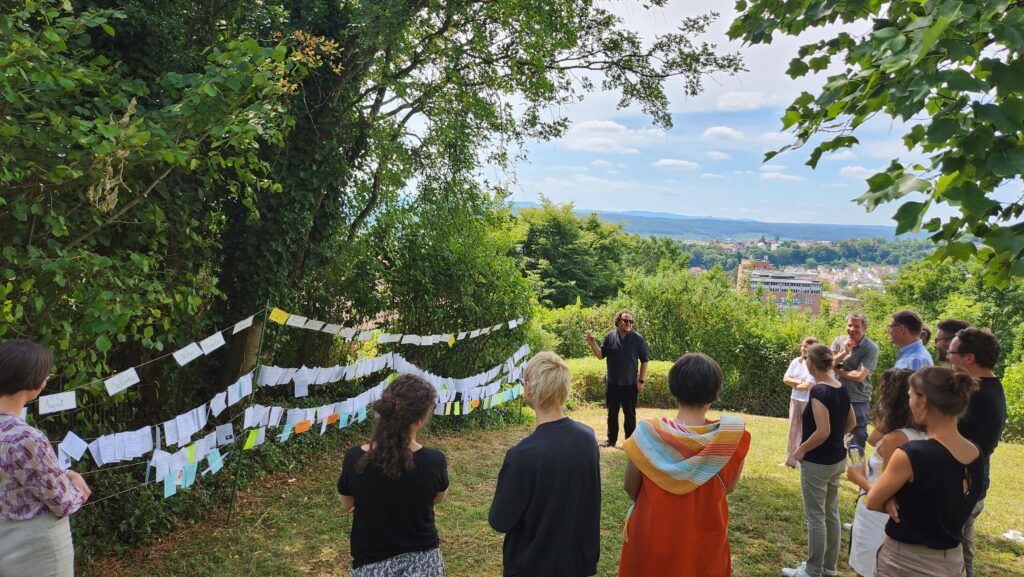 Das Bild zeigt eine Wäscheleine voller Zettel, die in einem Garten gespannt ist. Im Vordergrund sind Menschen zu sehen, die die Wäscheleine betrachten.