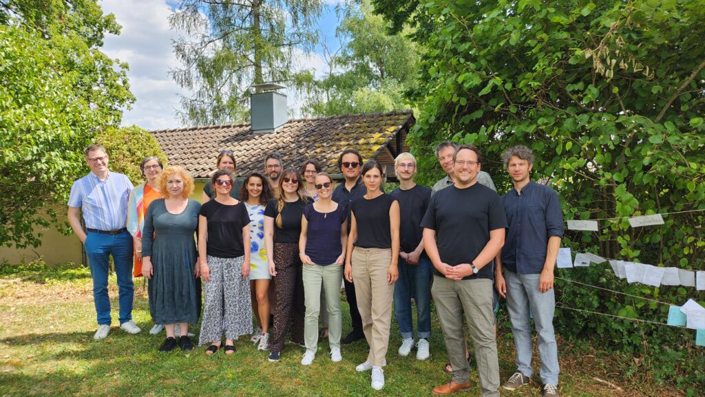 Das Bild zeigt eine Gruppe von 16 Personen, die vor einer Kulisse aus grünen Bäumen.